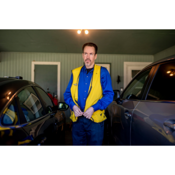 Photograph of Walmart cashier and self-checkout host Peter Naughton standing outside between two cars, wearing a blue button down and yellow vest Walmart uniform, looking into the distance. Naughton appears concerned.